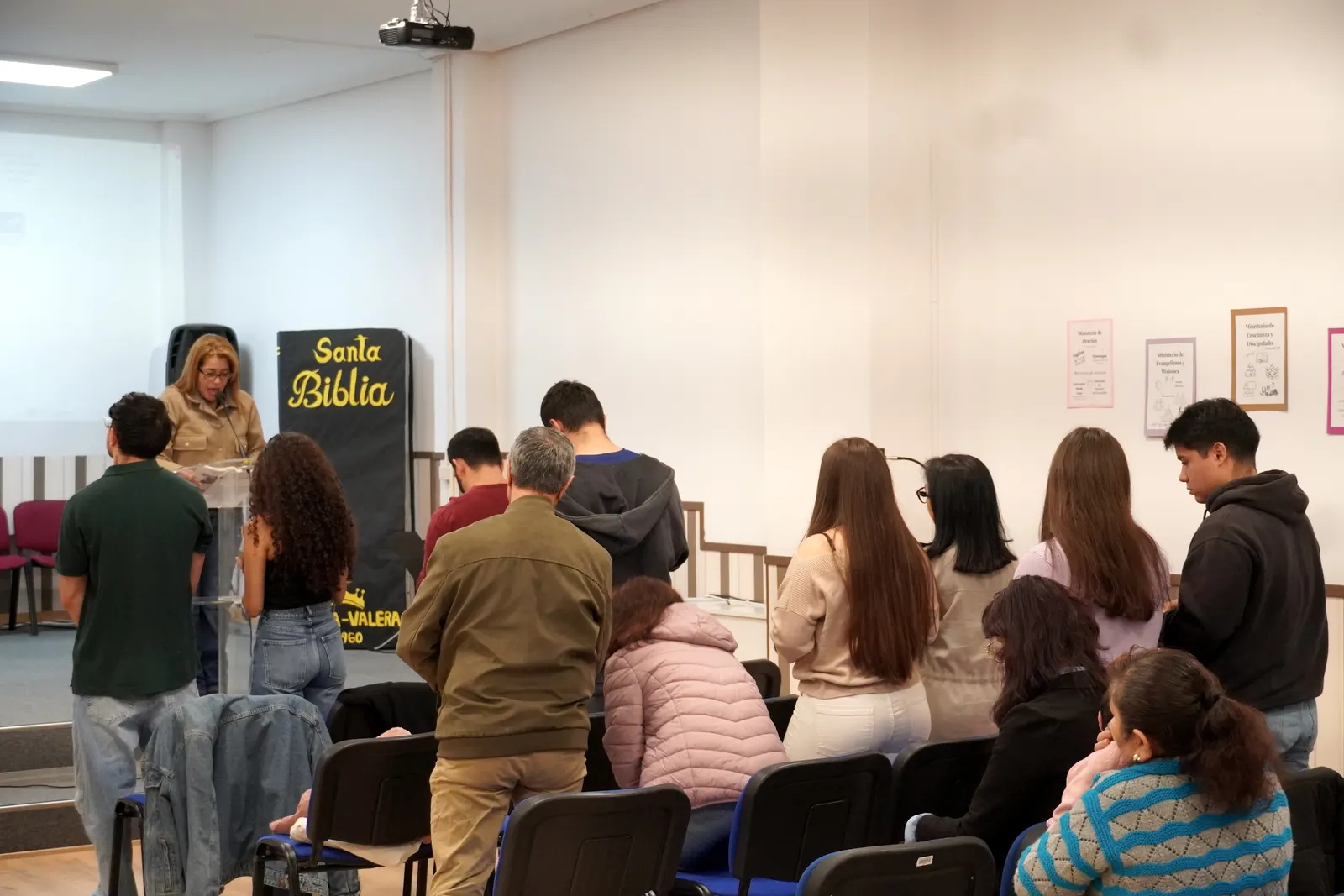 La congregación de pie durante el culto, con el cartel «Santa Biblia» al fondo